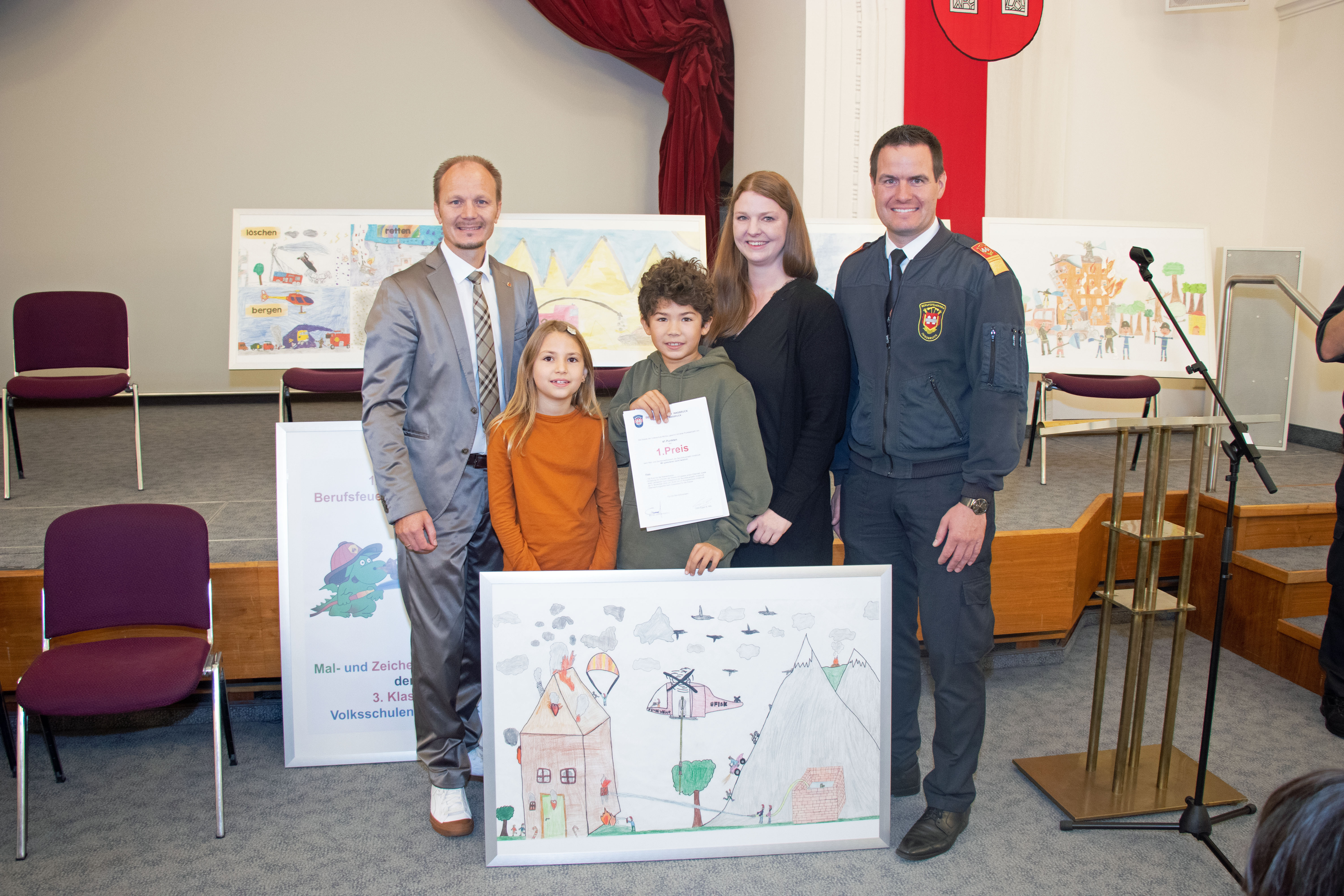 Die Volksschule Mühlau darf sich mit Vizebürgermeister Johannes Anzengruber (links) und Branddirektor Helmut Hager (rechts) über den ersten Preis freuen. Das ausgezeichnete Gemälde trägt den Titel "1-2-2 Zwergenfeuerwehr herbei". Die geschickte Verlegung des Einsatzortes in das Märchenland, die detaillierte Veranschaulichung der unterschiedlichen, an die vorliegende Notsituation angepassten Aufgaben der Feuerwehr im Einsatz, sowie der künstlerische Gesamteindruck konnten die Jury überzeugen. Als ersten Preis erhalten die Kinder 150€ für die Klassenkassa sowie eine Einladung zur Feuerwehr-Gala samt Überraschungspreis.